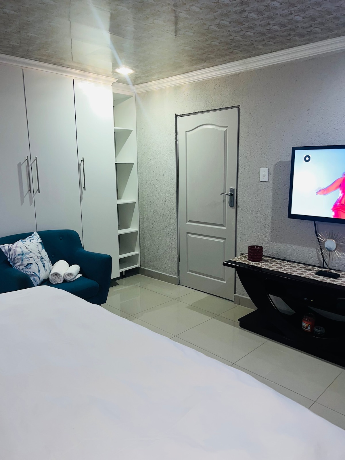 A compact living space includes a comfortable light blue armchair positioned beside a white wardrobe. A flat-screen TV is mounted on the wall, and a small black table supports a decorative item. Soft light reflects off polished tiles.