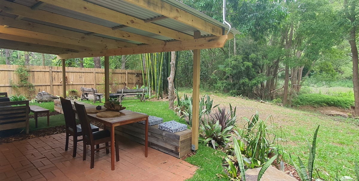 An outdoor dining area features a wooden table with seating for six under a shaded roof. Surrounding greenery includes various plants and trees, with a grassy area extending beyond the patio. A wooden bench provides additional seating, enhancing the natural setting.