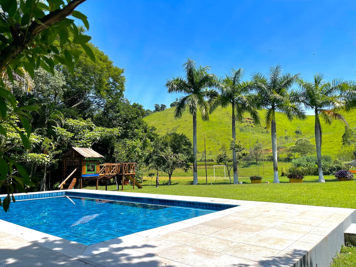 An outdoor area features a clear swimming pool surrounded by a stone patio. Lush green trees and rolling hills create a serene backdrop. A wooden playground is visible, along with a grassy field that includes a soccer goal, contributing to the spacious and inviting landscape.