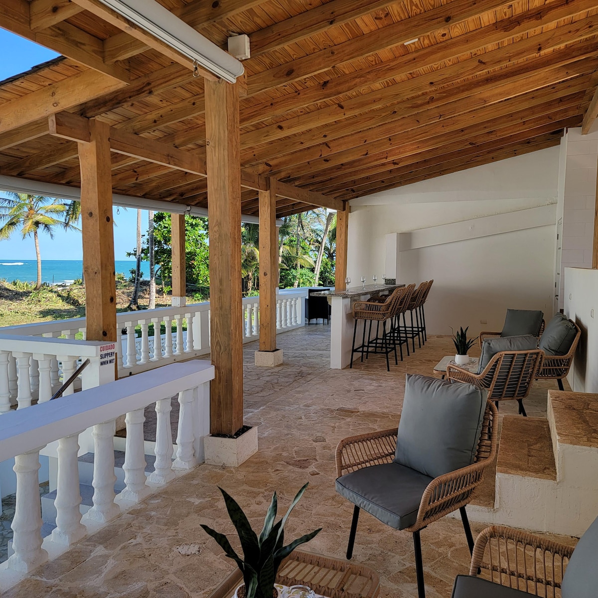An outdoor seating area is presented with wooden beams and a tiled flooring. Comfortable chairs and a small table are positioned beneath a shaded roof. Bar stools are arranged at a counter in the background, with views of a lush landscape and ocean visible in the distance.