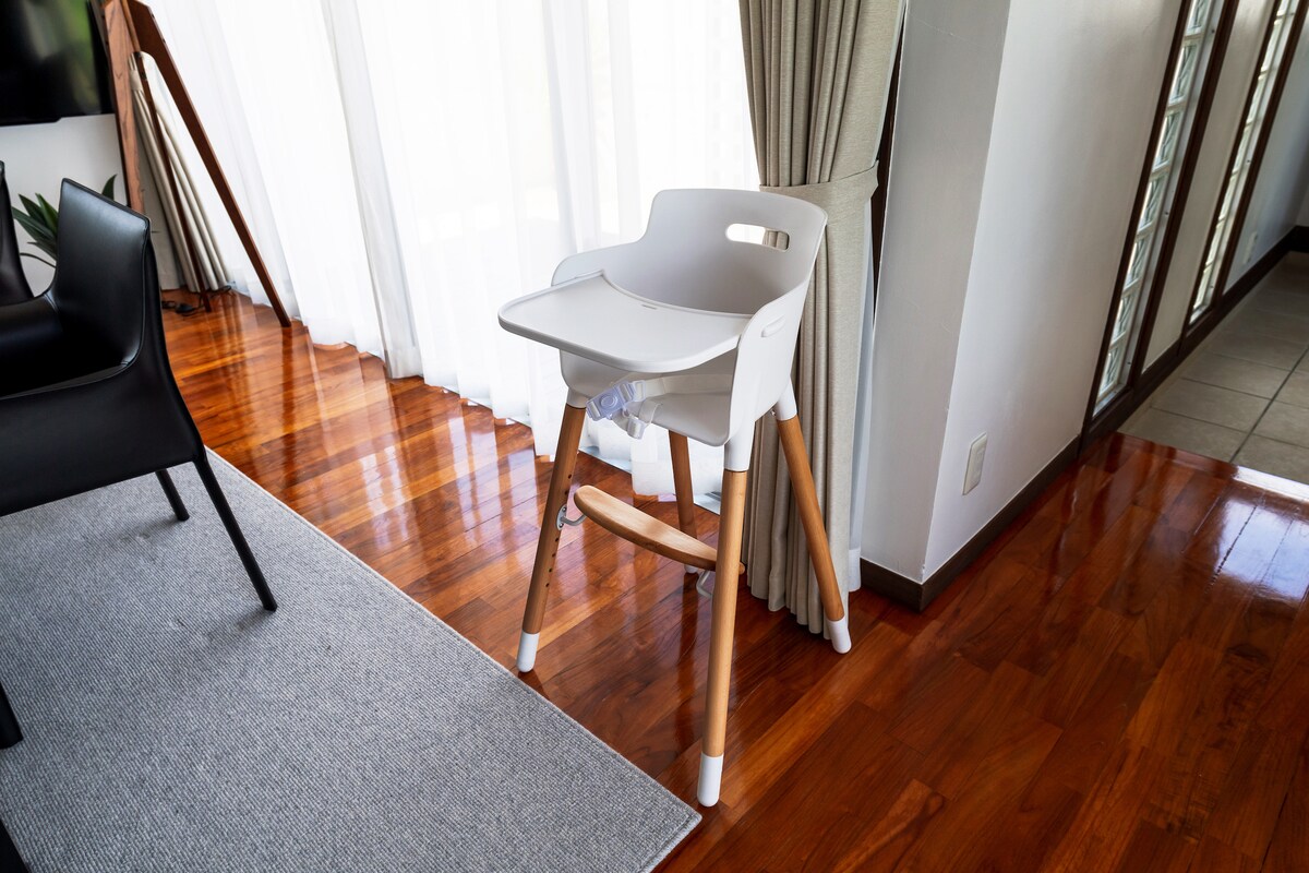 A high chair is positioned against a backdrop of large, sheer curtains allowing natural light to fill the space. Its minimalist design features a white seat with wooden legs, providing a functional option for families visiting the property.