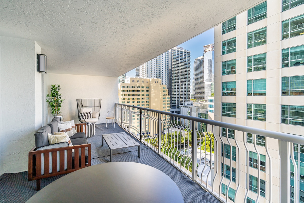 An outdoor patio features comfortable seating arrangements, including a sectional sofa with decorative cushions and a small table. The view captures a vibrant urban landscape filled with high-rise buildings and greenery, framed by a soft blue sky.