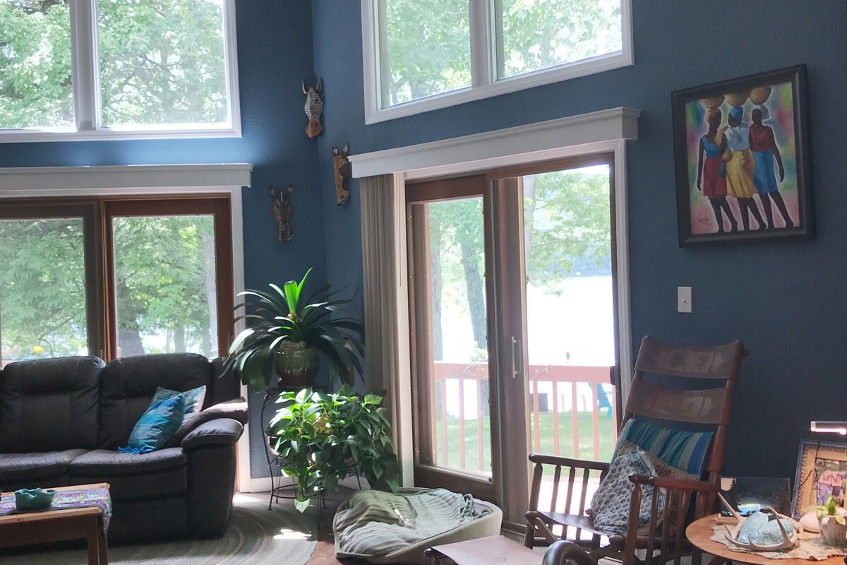 A spacious living area features large windows allowing natural light to illuminate the room, revealing a view of the waterfront. Comfortable seating is arranged around a coffee table, while a vibrant piece of wall art complements the decor. Lush greenery is present, adding a fresh touch.