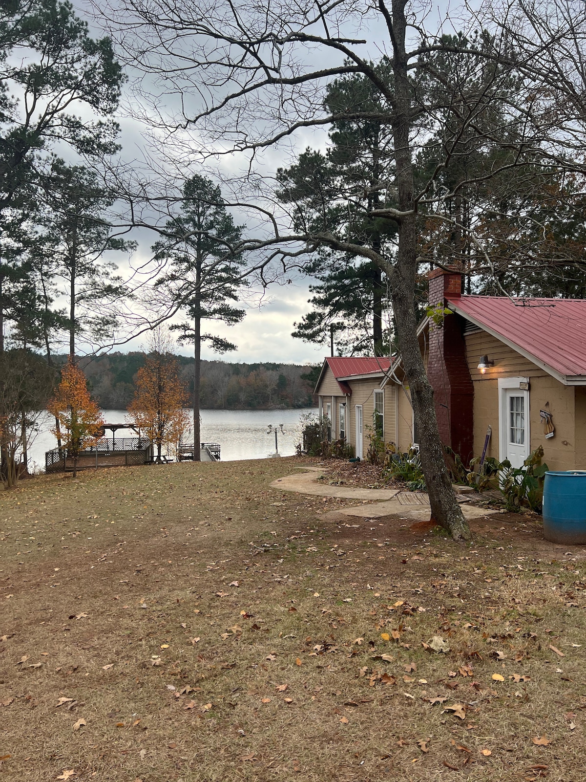 A peaceful view of a camp house is set against a serene lake. Tall trees frame the scene, while autumn foliage adds touches of color. A grassy area extends around the house, leading toward a private dock visible on the water's edge.