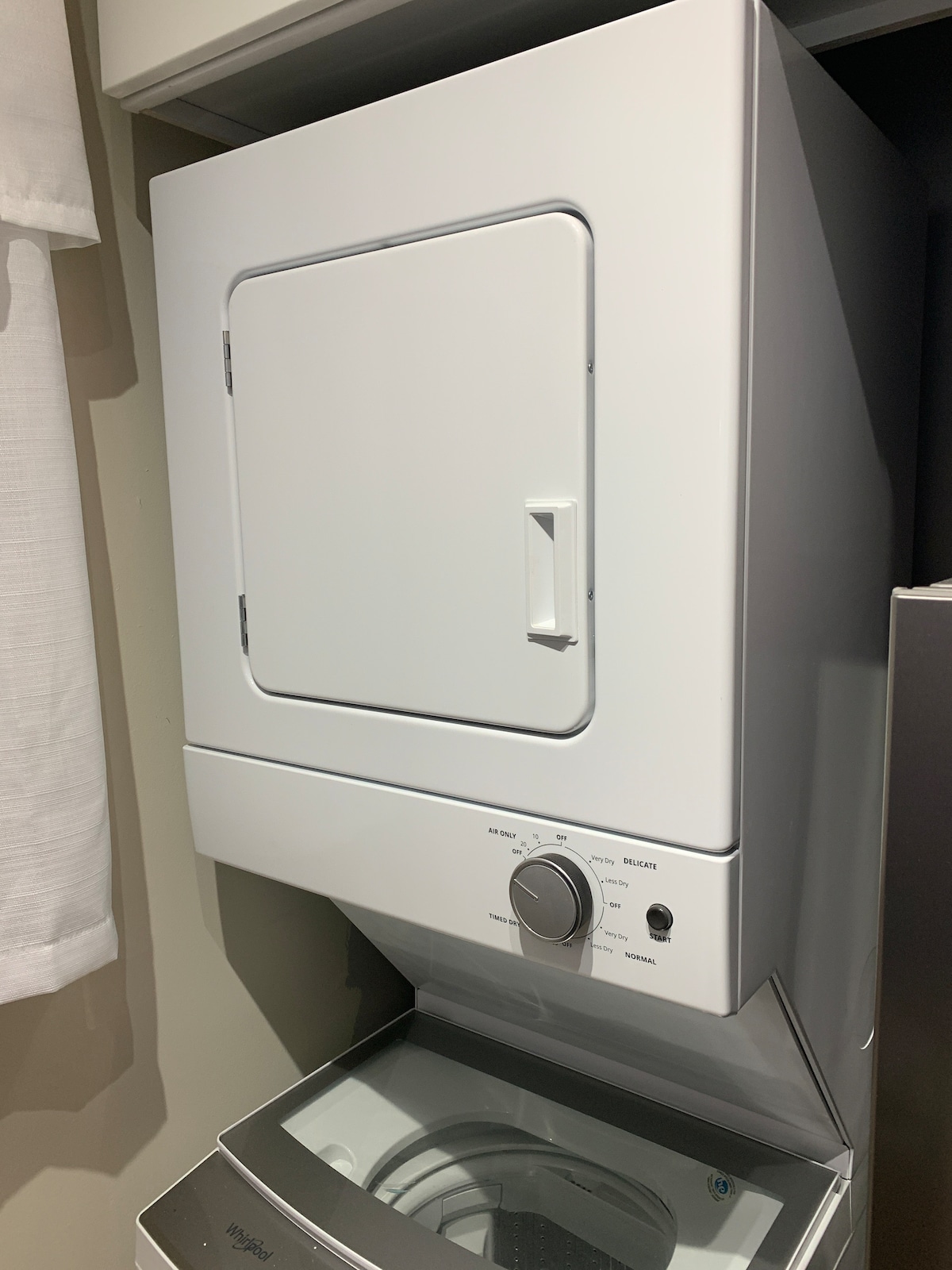 A stacked washer and dryer unit is positioned in a compact laundry area. The white exterior features a front-loading washer at the bottom and a dryer above, with a simple control dial and a door on the dryer for easy access.