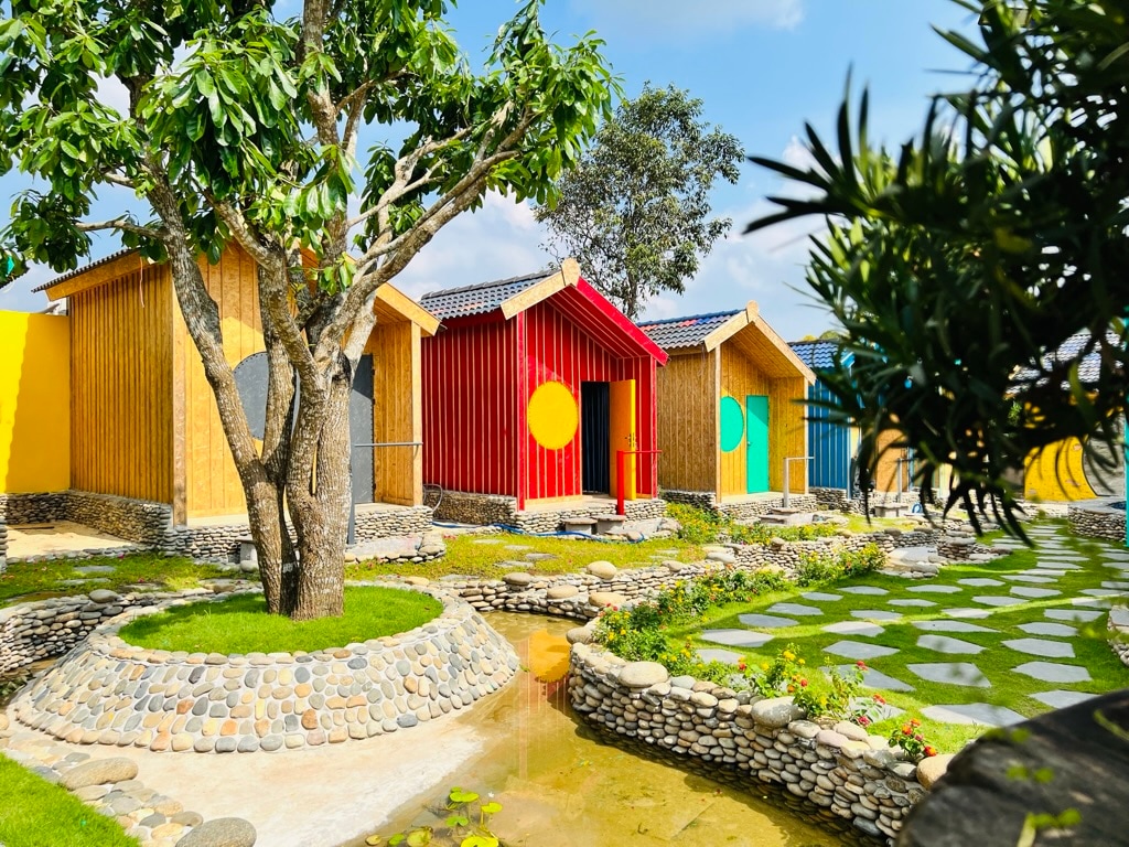 A cluster of rustic cabins is surrounded by landscaped greenery. Each cabin is painted in bright colors, with red, blue, and yellow accents, and is set against a clear blue sky. A winding stone pathway leads through the vibrant outdoor space.