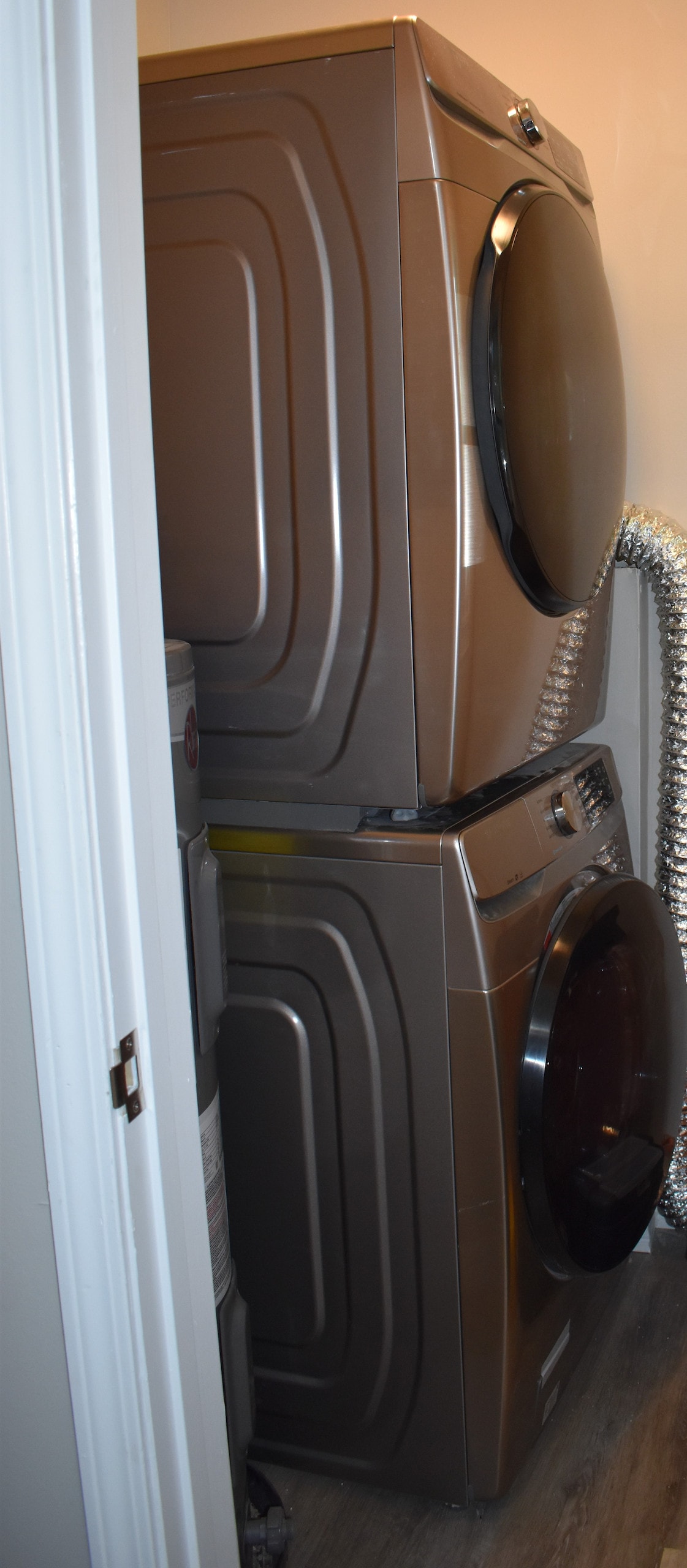 A stacked washer and dryer set in a compact space. The units are finished in a sleek metallic tone, positioned against a wall. A flexible duct can be seen attached to the dryer, facilitating ventilation.