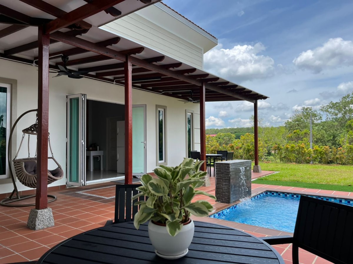 A modern outdoor seating area is visible, featuring a round table with black chairs and a potted plant at its center. A small swimming pool reflects the blue sky and green landscape, while a swing chair hangs from the covered porch, providing a relaxed atmosphere.