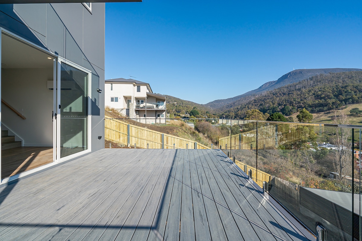A spacious outdoor deck is presented with a wooden surface and glass railing, showcasing views of rolling hills and Mount Wellington. The sunny sky adds brightness to the scene, while neighboring homes are visible on the opposite side of the valley.
