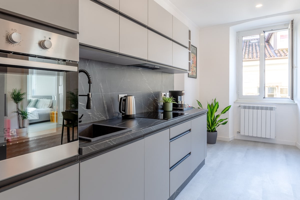 A modern kitchen is equipped with sleek cabinetry, a stovetop, and an oven. Counter space is complemented by a kettle and coffee machine. A large sink is included. A window allows natural light in, while a potted plant adds a touch of greenery to the space.