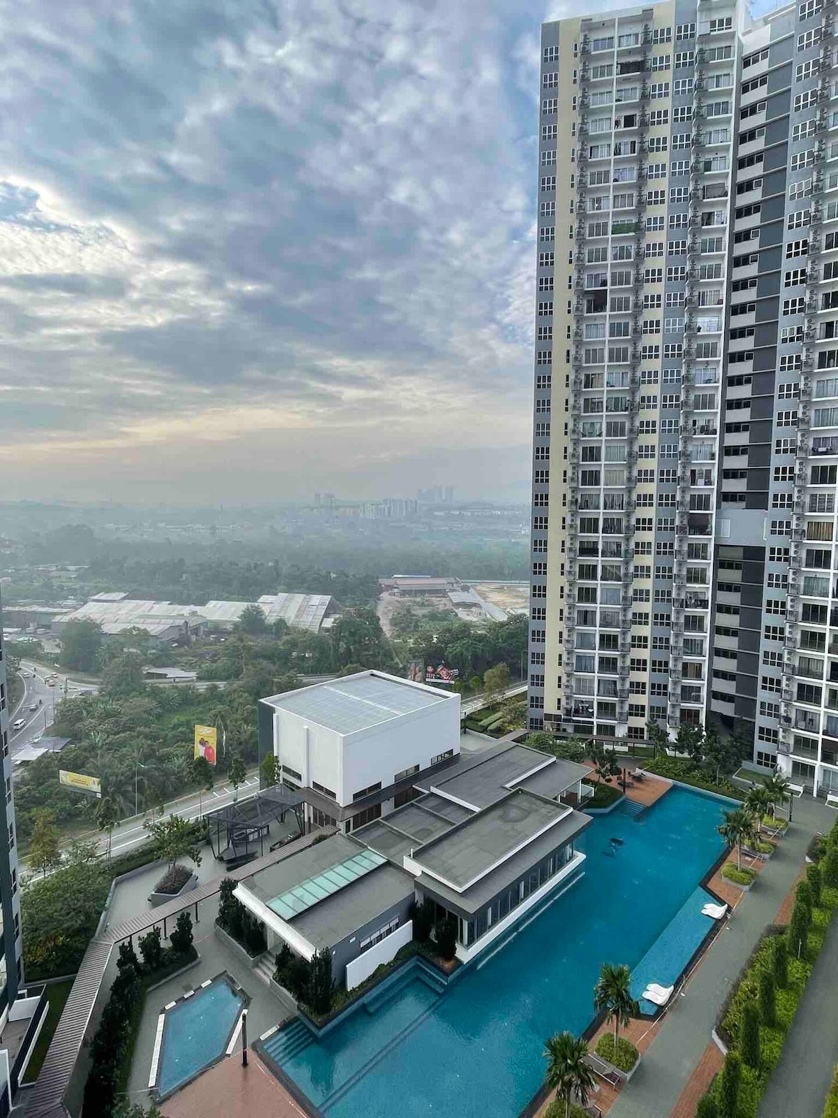 A cityscape view showcases a modern high-rise building alongside a landscaped outdoor area featuring an expansive infinity pool. Surrounding greenery and palm trees enhance the ambiance. The distant horizon reveals urban structures under a softly clouded sky.