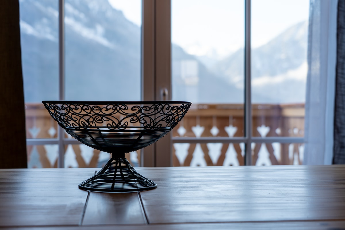 A metal fruit bowl is centered on a wooden table, featuring an intricate design. Large glass doors lead to a balcony, providing a view of distant mountains and natural light filling the room.
