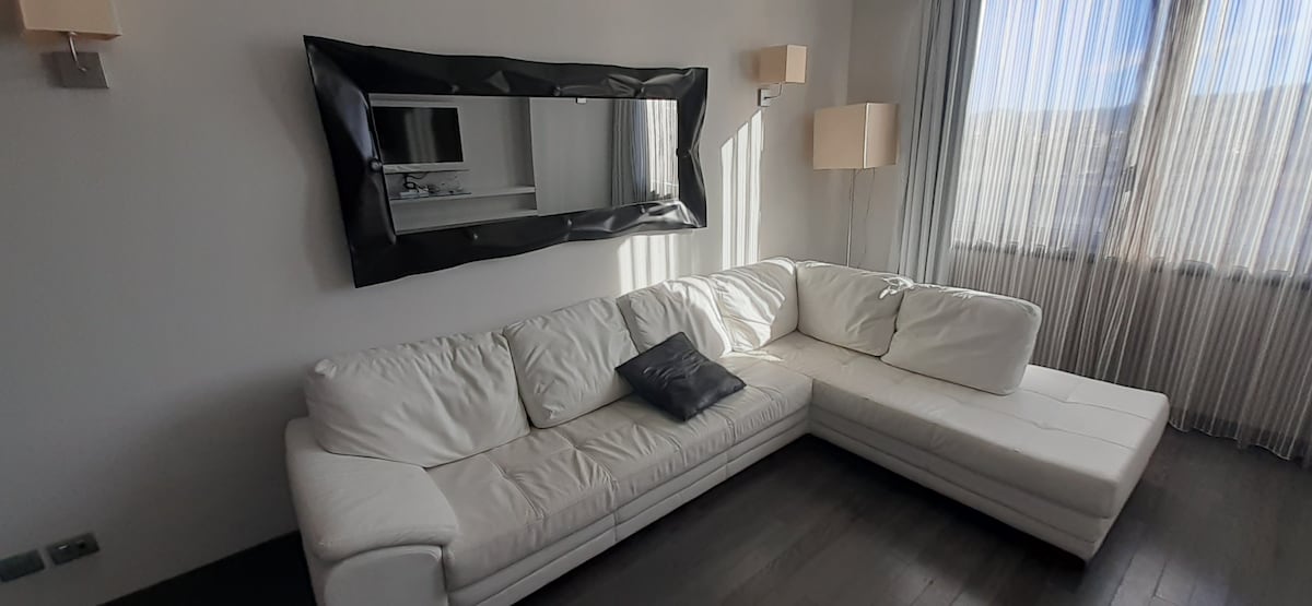 A spacious living area is furnished with a white sectional sofa featuring a modern design. Natural light filters through sheer curtains, highlighting a large wall mirror that reflects the room. Minimalistic lamps provide additional lighting.