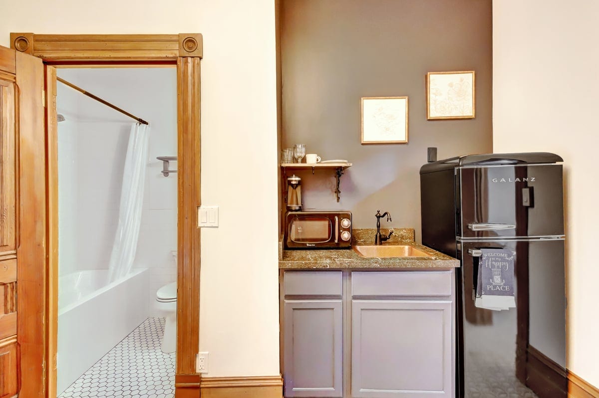 A compact kitchenette is visible, featuring a black retro fridge, a countertop sink, and a microwave. Above the counter, a shelf holds a coffee machine and essentials. A doorway leads to a bathroom with a bathtub and toilet, set against classic tile flooring.