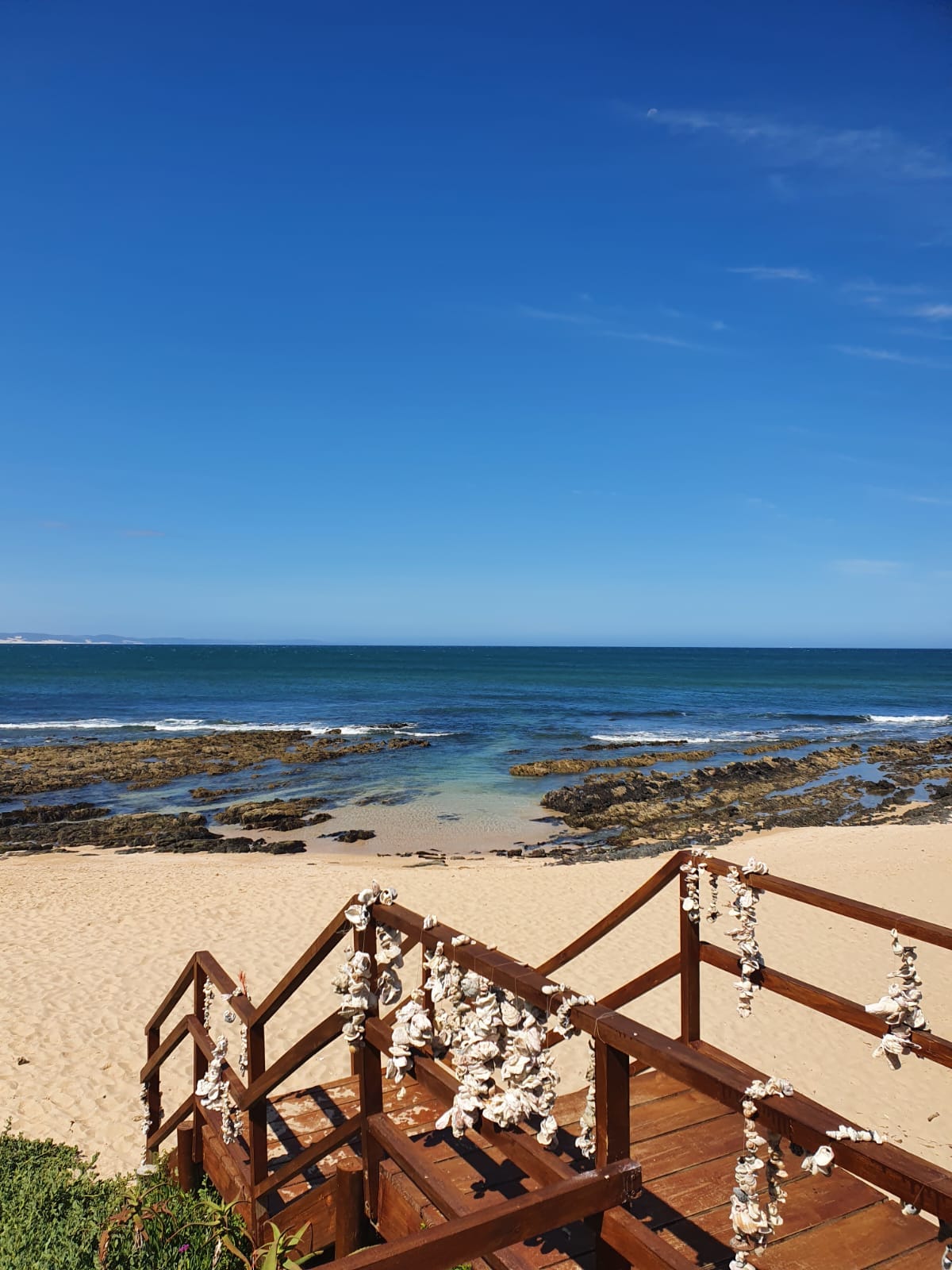 A sandy beach is visible, leading to calm blue waters scattered with rocks. A wooden railing, adorned with seashells, frames the view, enhancing the coastal scenery under a clear blue sky.