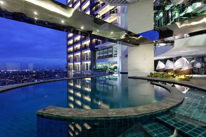 An illuminated pool area is viewed at dusk, surrounded by modern architecture. The pool's circular edge connects to a sleek walkway, reflecting the city skyline and ambient lights. Lush greenery is placed along the edges, adding a touch of nature to the contemporary setting.