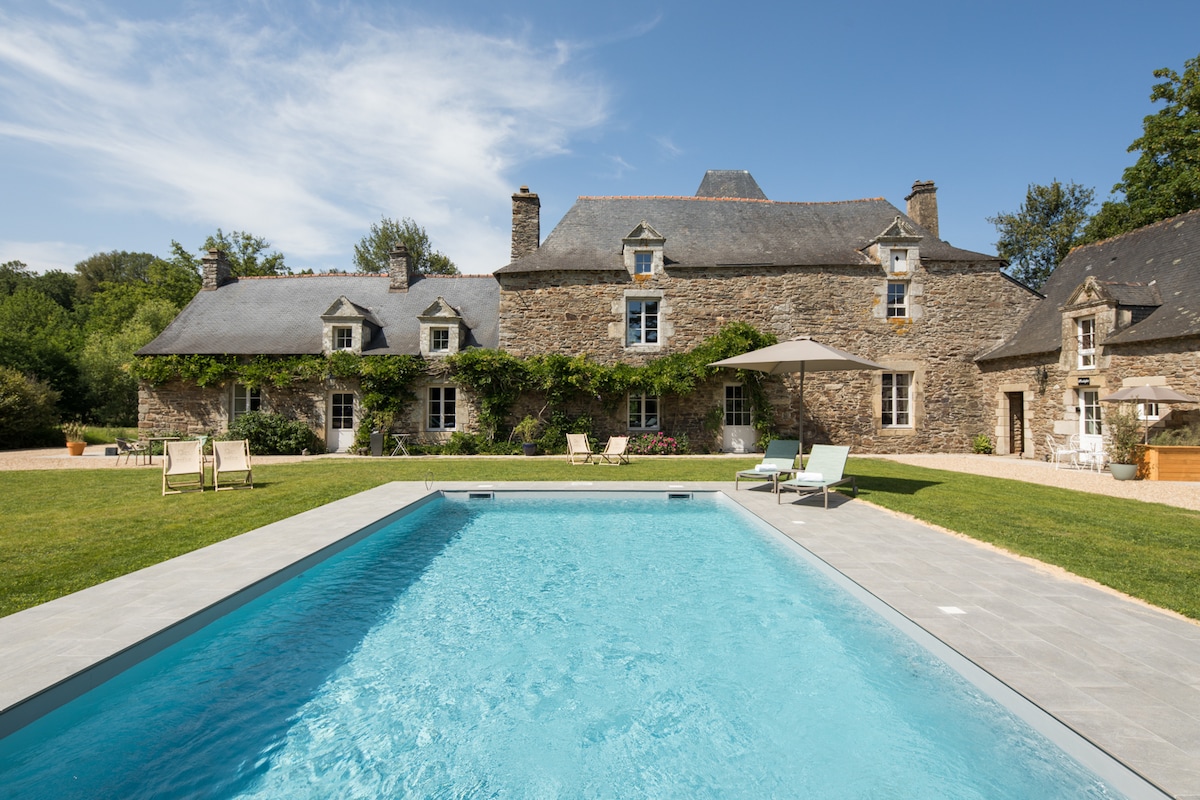 The exterior of a charming stone cottage is captured, showcasing a well-maintained swimming pool in the foreground. Lush greenery surrounds the property, complemented by sun loungers and a shaded area with an umbrella. The building features multiple windows, allowing natural light to illuminate the interior.