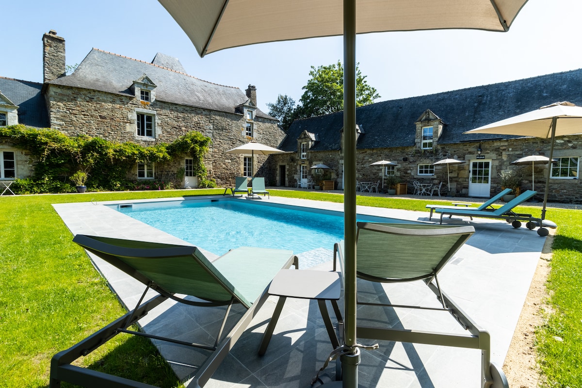 A refreshing swimming pool is framed by neatly manicured grass, with several lounge chairs and umbrellas providing shade. The historic stone buildings of the cottage are visible in the background, surrounded by lush greenery, creating a serene outdoor setting.