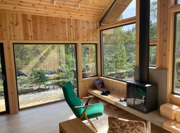 A bright living area is revealed, featuring large windows that provide views of the surrounding greenery. A modern wood stove is situated on a concrete base, complemented by a wooden coffee table and a green armchair. Natural light enhances the warm wooden interior.