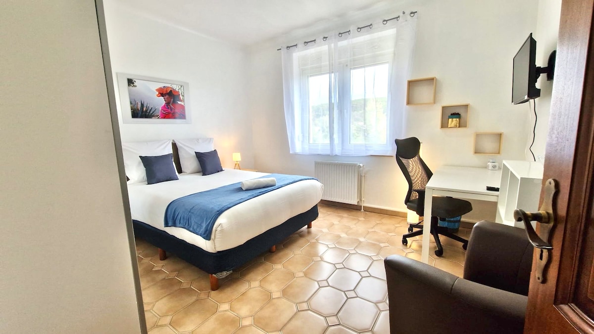 A bright room features a spacious bed dressed in crisp white linens with two accent pillows. A desk area includes a comfortable office chair and a wall-mounted smart TV. Natural light filters through the double-glazed window, and a cozy armchair complements the inviting ambiance.