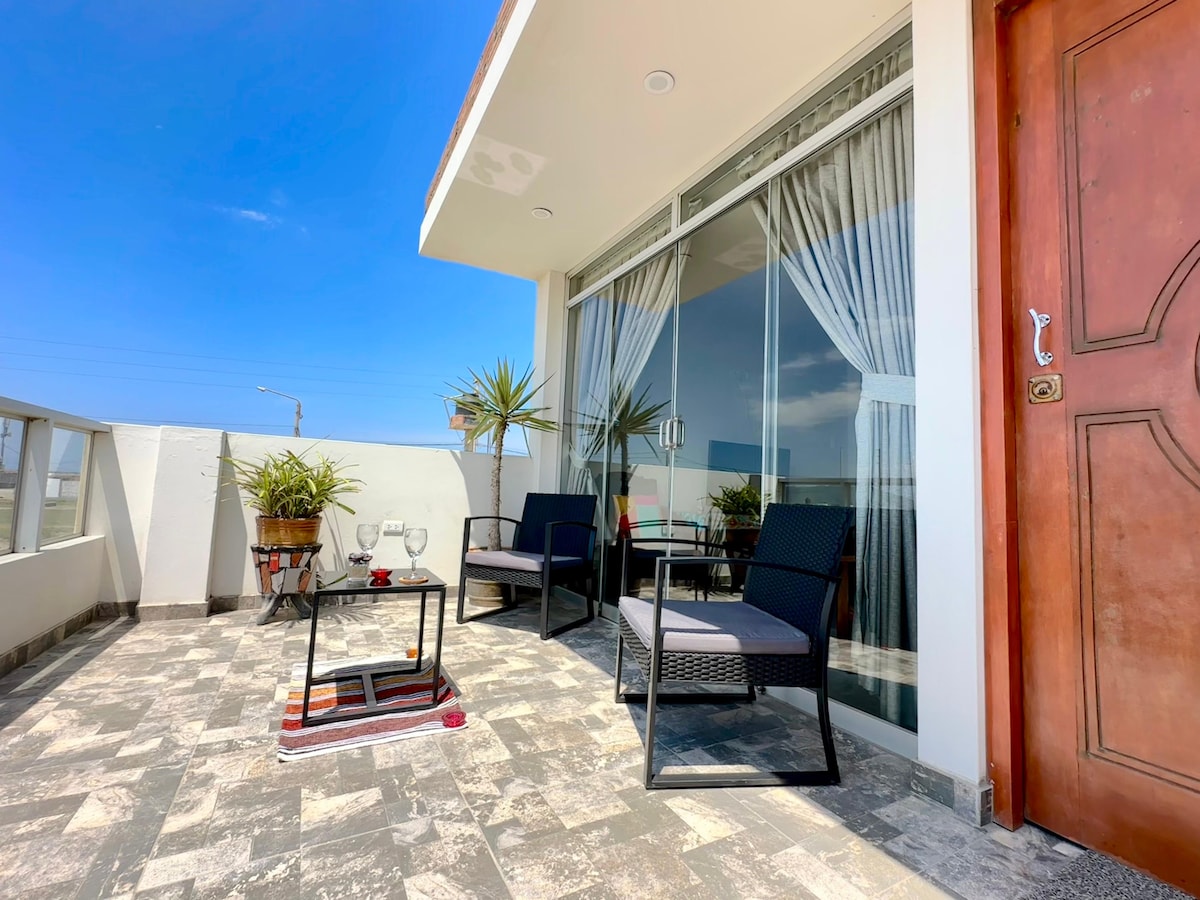 A private terrace is visible, featuring modern outdoor furniture including two black wicker chairs and a small glass table. A plant adds a touch of greenery, and large glass doors provide an open view, allowing natural light to fill the space.