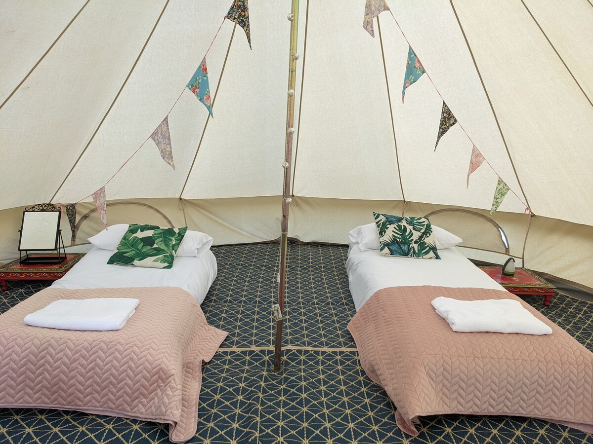 Inside a spacious bell tent, two single futon beds are neatly arranged, each with soft bedding and towels. Decorative cushions add a touch of comfort, while bunting and fairy lights enhance the inviting atmosphere. A mirror is positioned to the side, reflecting the cozy interior.