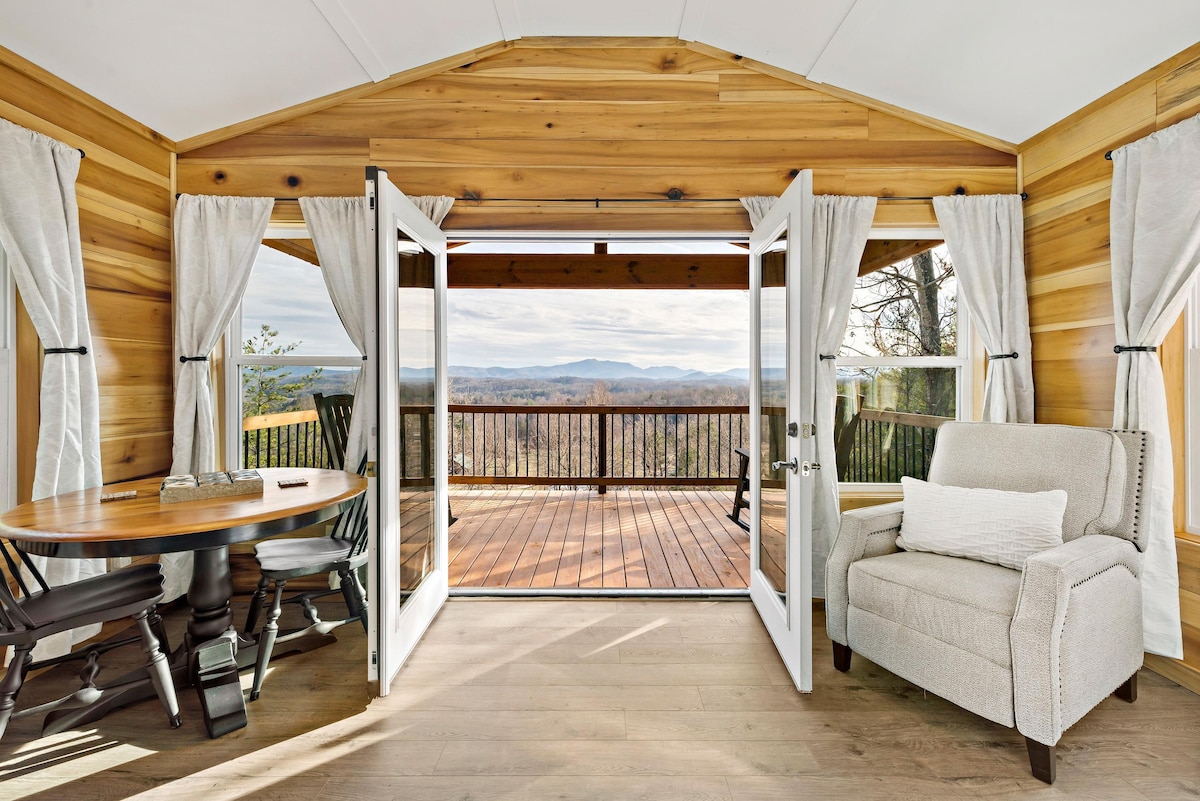 A spacious interior features warm wood paneling and opens to a large deck through French doors. A round dining table is paired with elegant dining chairs, while a comfortable armchair sits nearby. Scenic mountain views are visible through the doors.
