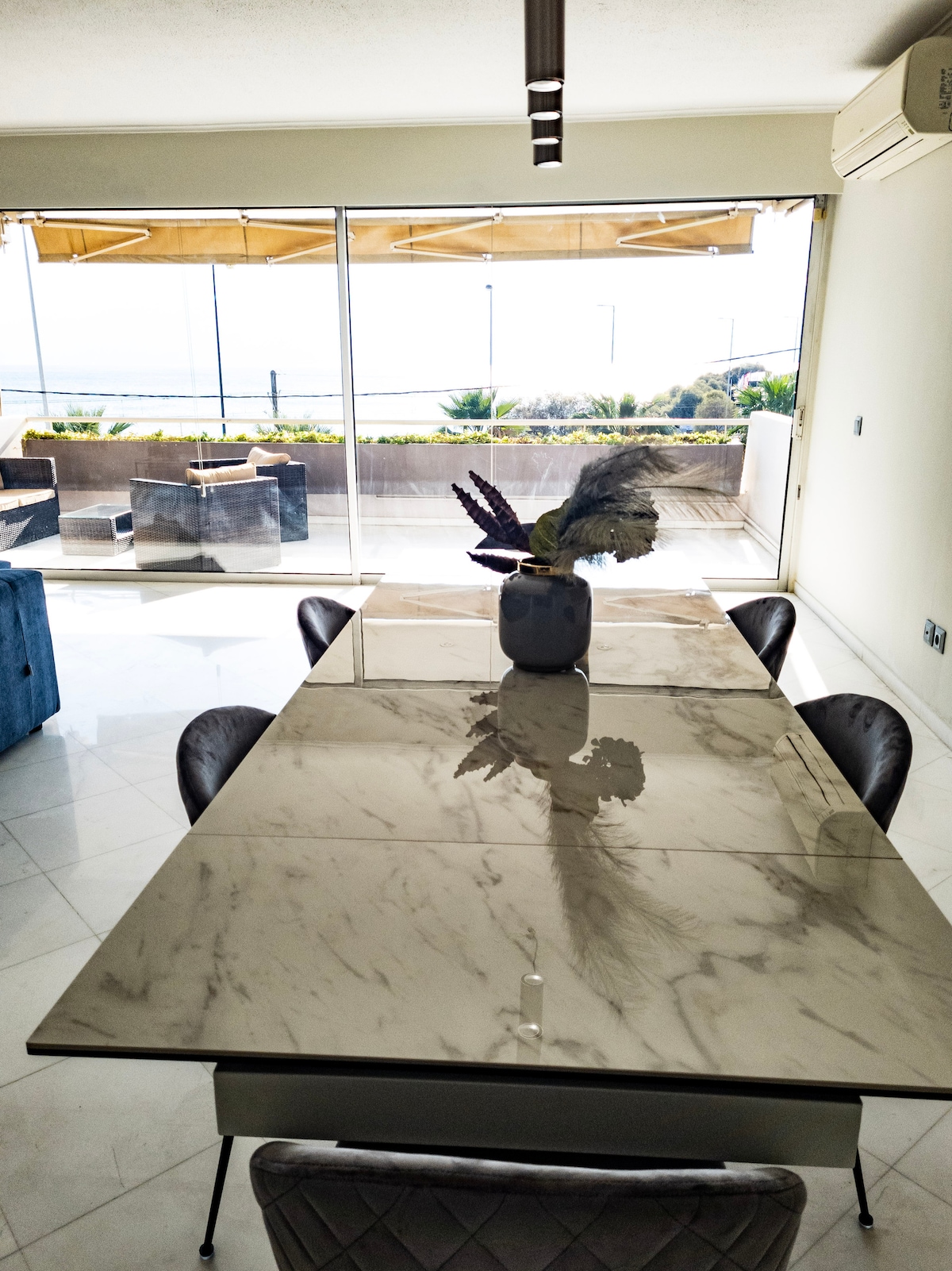 A modern dining area features a sleek marble table surrounded by four chairs. A vase with decorative foliage is positioned at the center of the table. Through expansive glass doors, a patio is visible, adorned with outdoor seating and offering views of the beach.