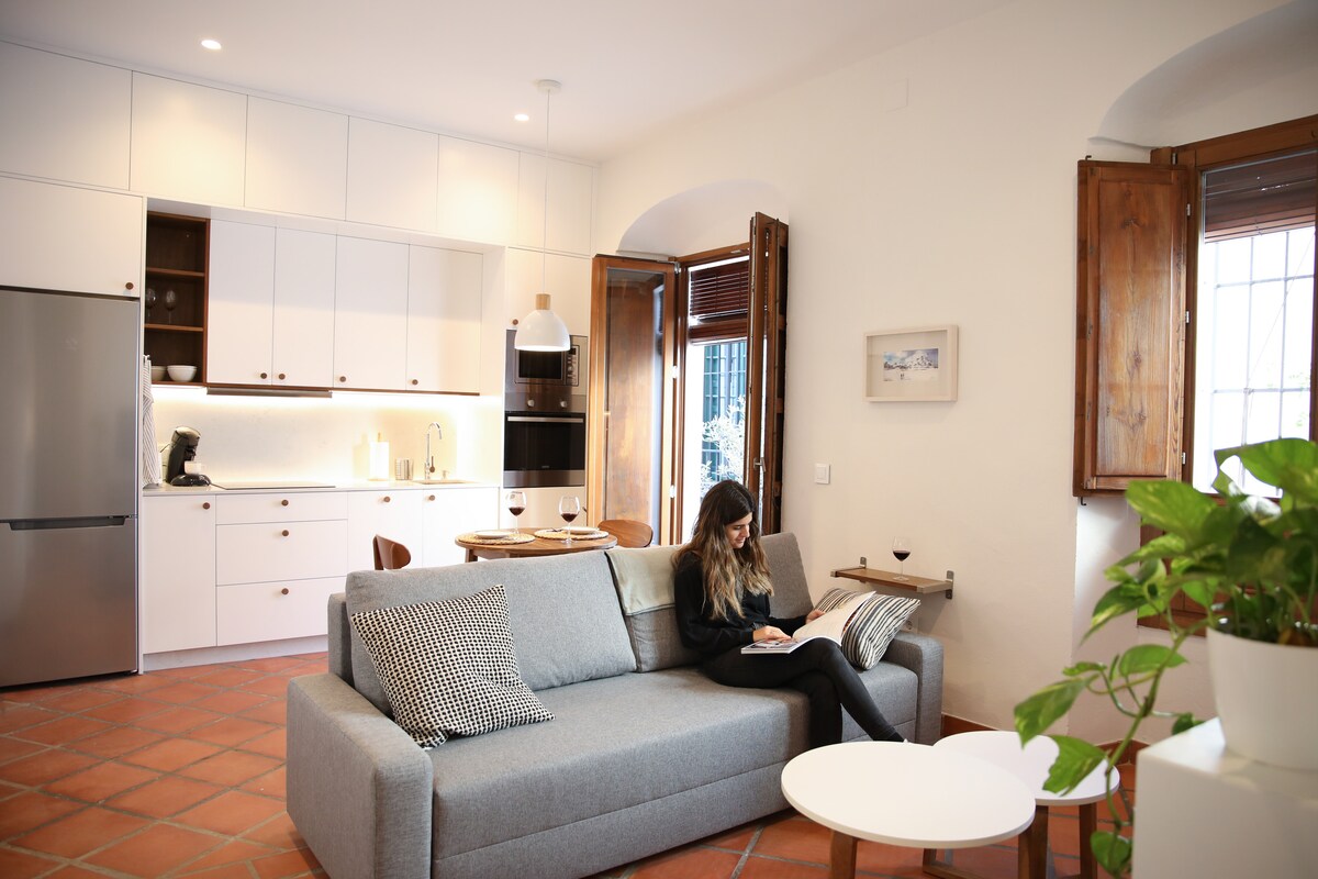A modern living area showcases a comfortable gray sofa paired with decorative cushions. Light fills the space from the open wooden windows, while the kitchen features sleek cabinetry and modern appliances. A small white coffee table enhances the inviting layout of the room.