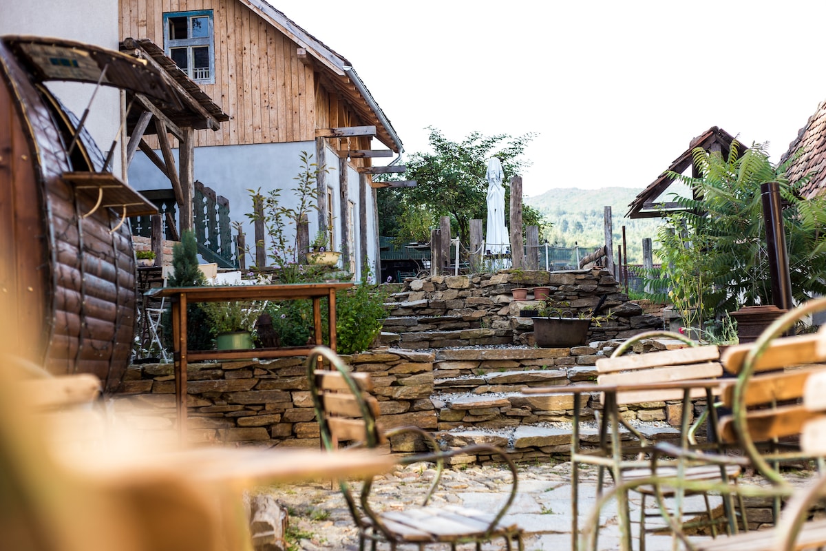 A peaceful outdoor area features stone pathways leading to lush greenery and rustic buildings. Wooden tables and chairs are arranged for outdoor seating, framed by natural stone accents and potted plants. The backdrop includes a quaint wooden structure and a shaded umbrella.