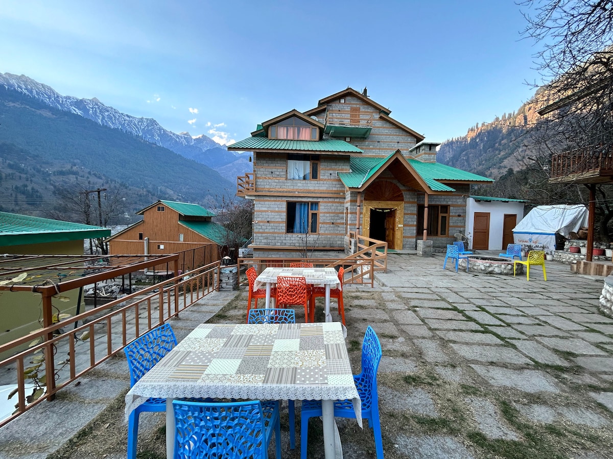 An inviting outdoor area showcases colorful tables and chairs set upon stone paving. The wooden structure of the cottage is visible, nestled against a backdrop of majestic mountains. Clear skies illuminate the setting, highlighting the natural beauty of the surrounding landscape.