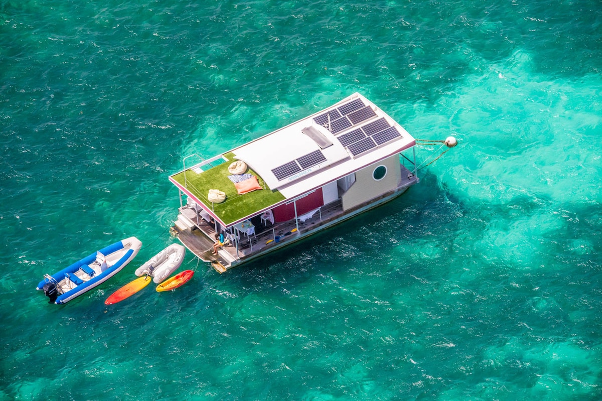 An aerial view showcases a floating villa surrounded by vibrant turquoise waters. Solar panels are installed on the roof, while two kayaks rest beside the docked vessel. The outdoor space includes a green terrace with lounge chairs, providing an inviting area for relaxation.