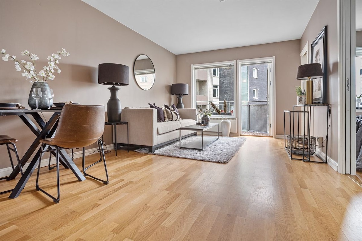 A bright living space features wooden flooring, complemented by a light rug. A neutral sofa is arranged with decorative cushions, across from a dining table set for four. Large glass doors provide views of a balcony, enhancing the openness of the area.