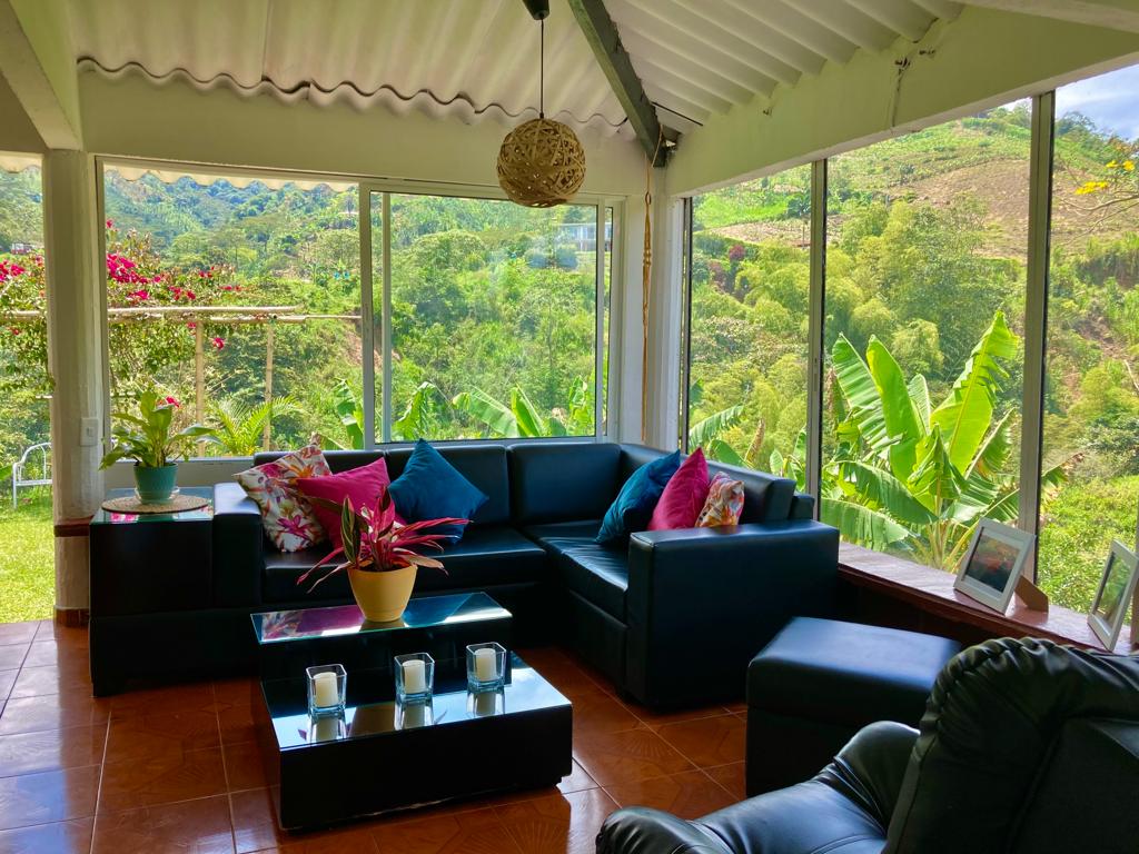 A spacious living area features large windows allowing natural light to fill the space, complemented by views of the surrounding greenery. A black sectional sofa with colorful cushions offers comfortable seating, while a coffee table is set with decorative candles. Vibrant plants add a touch of nature.
