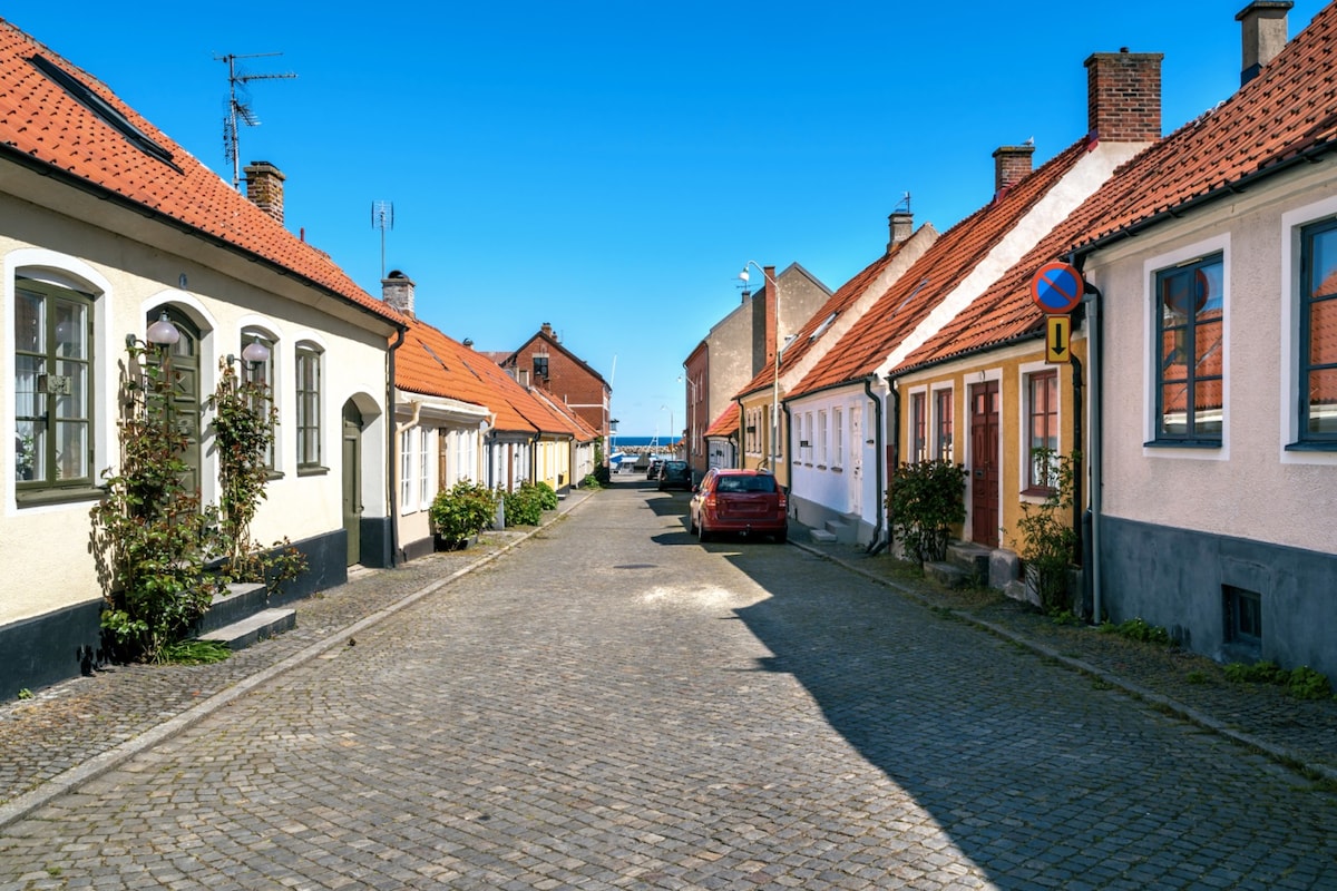 A tranquil street scene features charming houses with red roofs and cobblestone pavement. Buildings in various pastel colors are lined along the street, with windows framed by greenery. A glimpse of the sea can be seen at the end of the road, bathed in bright sunlight.