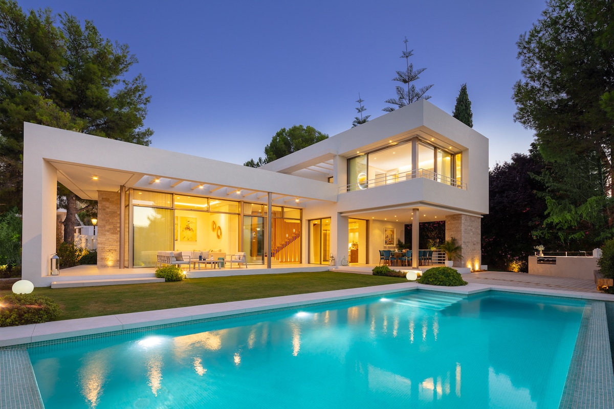 A modern home is presented, featuring large glass windows that create a seamless flow between indoor and outdoor spaces. The inviting turquoise pool reflects the surrounding greenery, while softly lit landscaping enhances the evening ambiance. A staircase leads to the upper level, surrounded by lush trees.