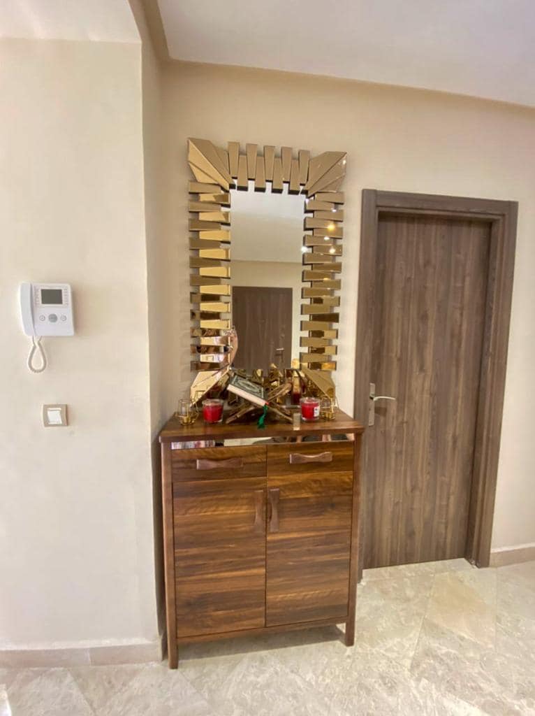 An elegant wooden cabinet with two drawers stands against a neutral wall. A decorative mirror with a sunburst design is mounted above the cabinet. Small items and decorative candles are placed on top, adding a touch of style to the entryway.