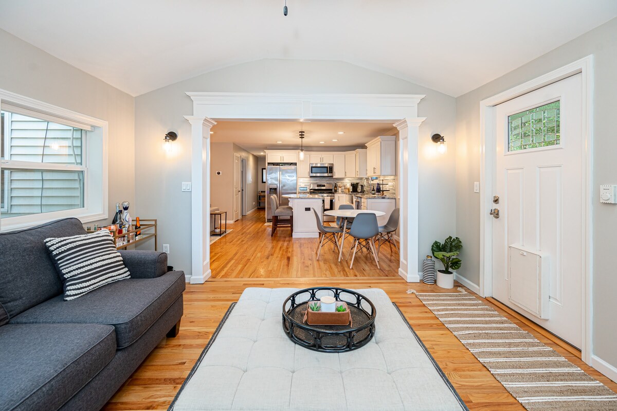 The living room features a large gray couch with accent pillows, complemented by a round coffee table at the center. Light wood flooring leads to an open space that connects to a well-equipped kitchen. Natural light enters through windows, enhancing the inviting ambiance.