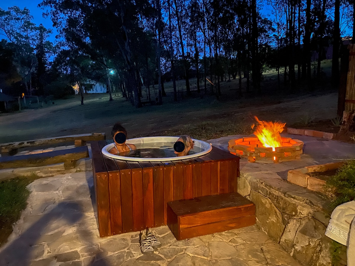 A rustic hot tub, crafted from wood, is positioned beside a circular fire pit with a lively flame. The evening setting creates an inviting ambiance, with trees softly illuminated in the background. Stone pathways connect these outdoor relaxation areas, enhancing the natural surroundings.