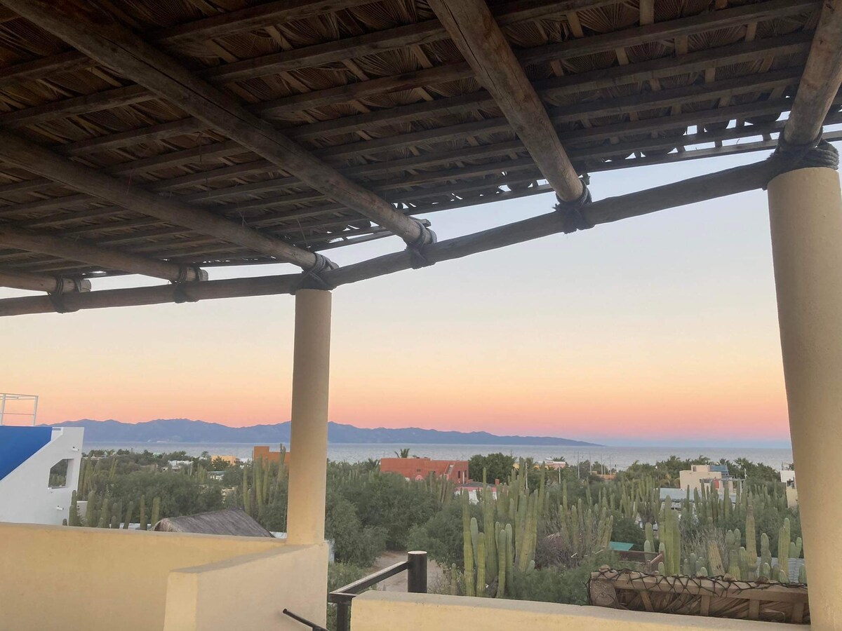 The view from a shaded outdoor area features a vibrant sunrise or sunset, with soft pastel colors blending in the sky. The foreground is framed by a rustic wooden roof, while the landscape includes a mix of cacti and distant mountains leading to the horizon.