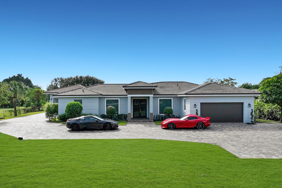 A modern home is presented with a well-kept lawn and circular driveway. Two vehicles are parked in front: a black car and a red sports car. The neatly designed exterior features large windows, complemented by manicured shrubbery and trees surrounding the property.