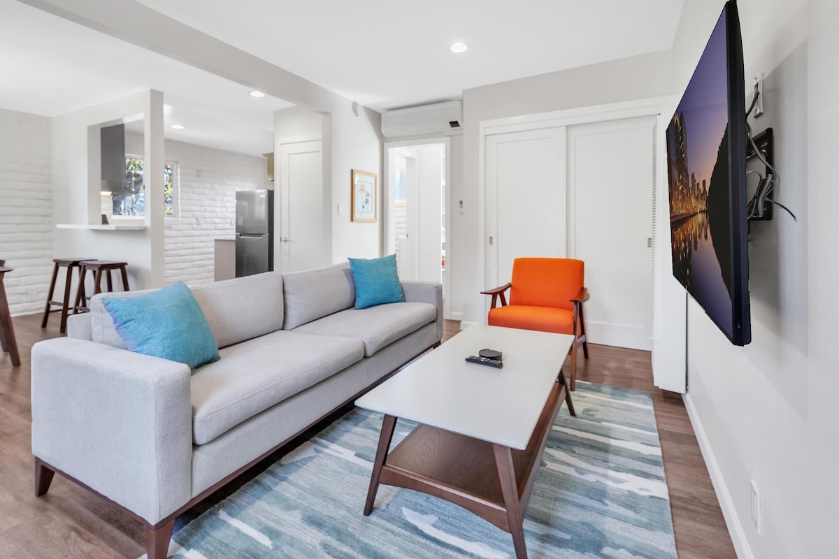 A bright living area features a light gray sofa adorned with blue cushions, complementing an orange chair. A wooden coffee table sits centrally, and a wall-mounted TV is visible. Natural light fills the space through nearby doors leading to the outdoor area.