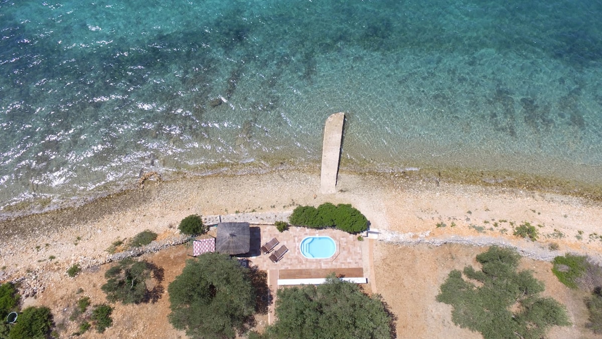 An aerial view captures a coastal retreat featuring a small pool surrounded by a tiled patio. Sun loungers offer space for relaxation under the sun, complemented by a stony shoreline and clear turquoise waters. Lush greenery borders the area, enhancing the natural landscape.