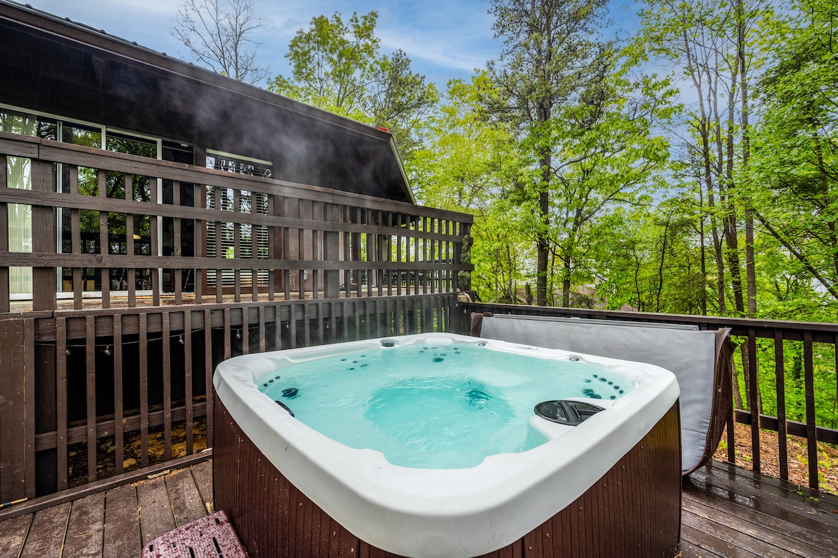 A hot tub is situated on a spacious wooden deck surrounded by vibrant green trees. Steam rises gently from the bubbling water, creating a serene ambiance. The cabin's structure is partially visible in the background, providing a natural feel to the private outdoor space.