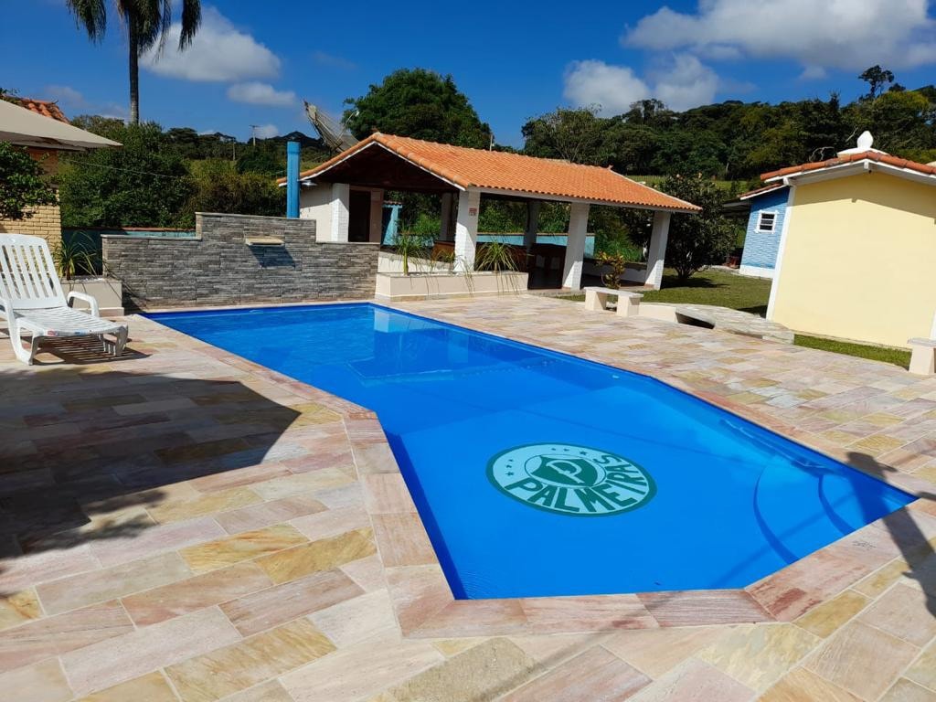A rectangular swimming pool is surrounded by a stone patio, featuring lounge chairs positioned at the edge. The pool is vibrant blue with a logo at the center. Nearby, a barbecue area and several buildings are visible, framed by greenery and a clear blue sky.