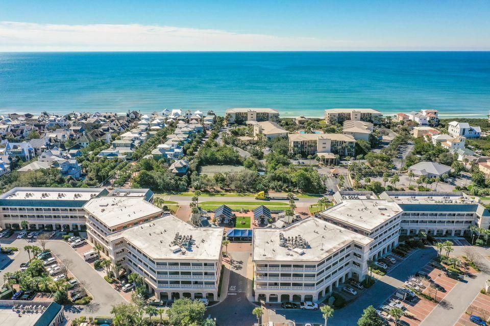 An aerial view of a coastal area is presented, featuring a cluster of condo buildings arranged in a U-shape. The ocean is visible in the background, with a serene shoreline lined by residential properties. Lush greenery and pathways are interspersed throughout the landscape.