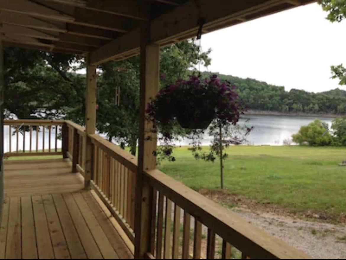 A wooden deck extends along the cottage, providing a serene view of Table Rock Lake. Lush greenery borders the water, while a hanging planter of purple flowers adds a touch of color. The scene invites relaxation and appreciation for the tranquil surroundings.