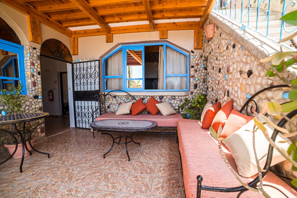 An outdoor seating area features a large stone wall with decorative elements. Comfortable seating is provided by a long bench with colorful cushions, accompanied by a round table. Natural light filters in through the blue-framed windows, enhancing the inviting atmosphere.