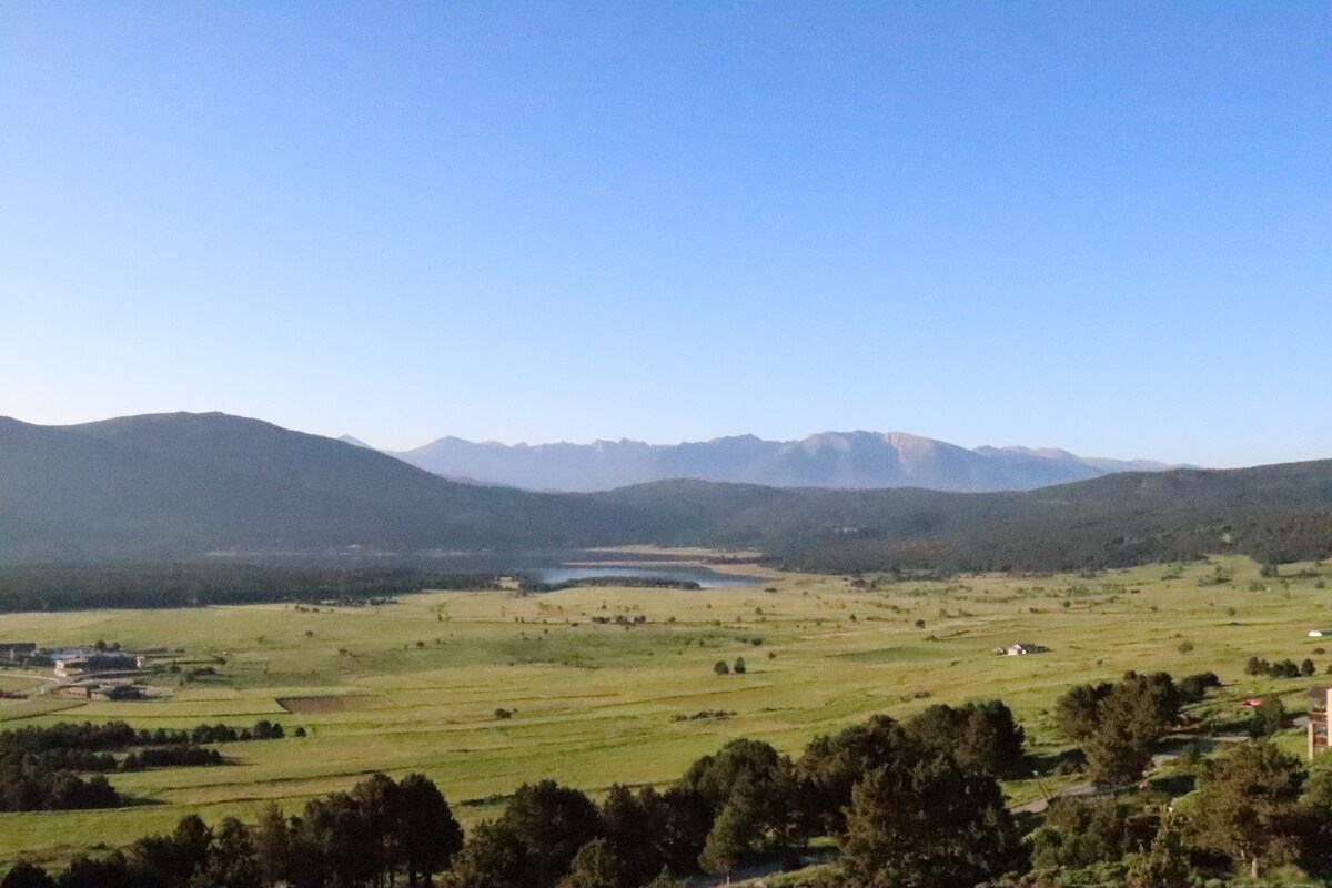 A wide landscape view showcases a serene valley with rolling hills and distant mountains. A lake can be seen calmly reflecting the sky. The foreground features a mix of greenery and open fields, enhancing the natural beauty of the surrounding area.