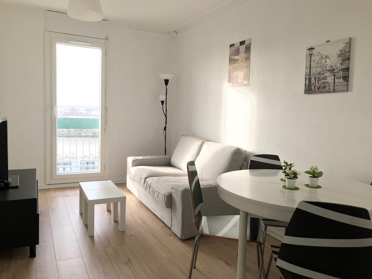 A bright living area features a light-colored sofa and a small coffee table, complemented by a round dining table surrounded by striped chairs. Two framed images adorn the walls, and natural light fills the space through a large window offering views beyond.